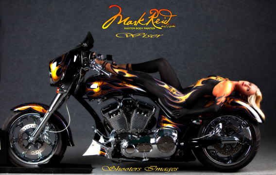 blonde woman in full boday paint featuring flames on a black background laying on her back on a motorcycle with the same design